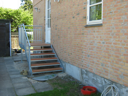 HOVEDTRAPPE med hårdttræ-trin og stålrør- håndliste. Hovedtrappen`s repos monteres med stålbolte i husmur under hoveddør (eller som her, på bærende stålsøjle) og trappen fastboltes på 2 punktfundamenter, et for bærende søjle yderst ved reposhjørne + punktfundament ved start af trappetrin. Hovedtrappen`s punktfundamenter kan etableres i færdig nivau, eller som her, under færdig belægnings-overflade. 
